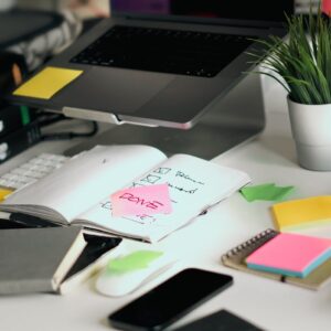 A cluttered office desk featuring a laptop, notebooks, and checklists amid colorful sticky notes.